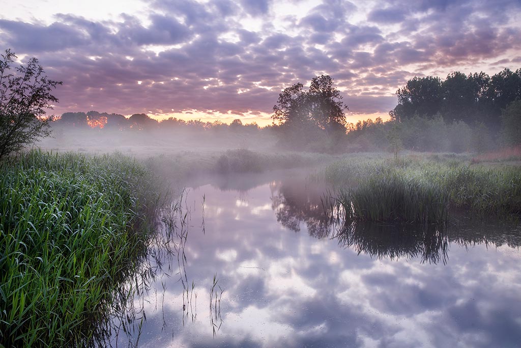 Op een mistige, rustige ochtend reflecteert het gladde water perfect de kleurrijke wolkenlucht.